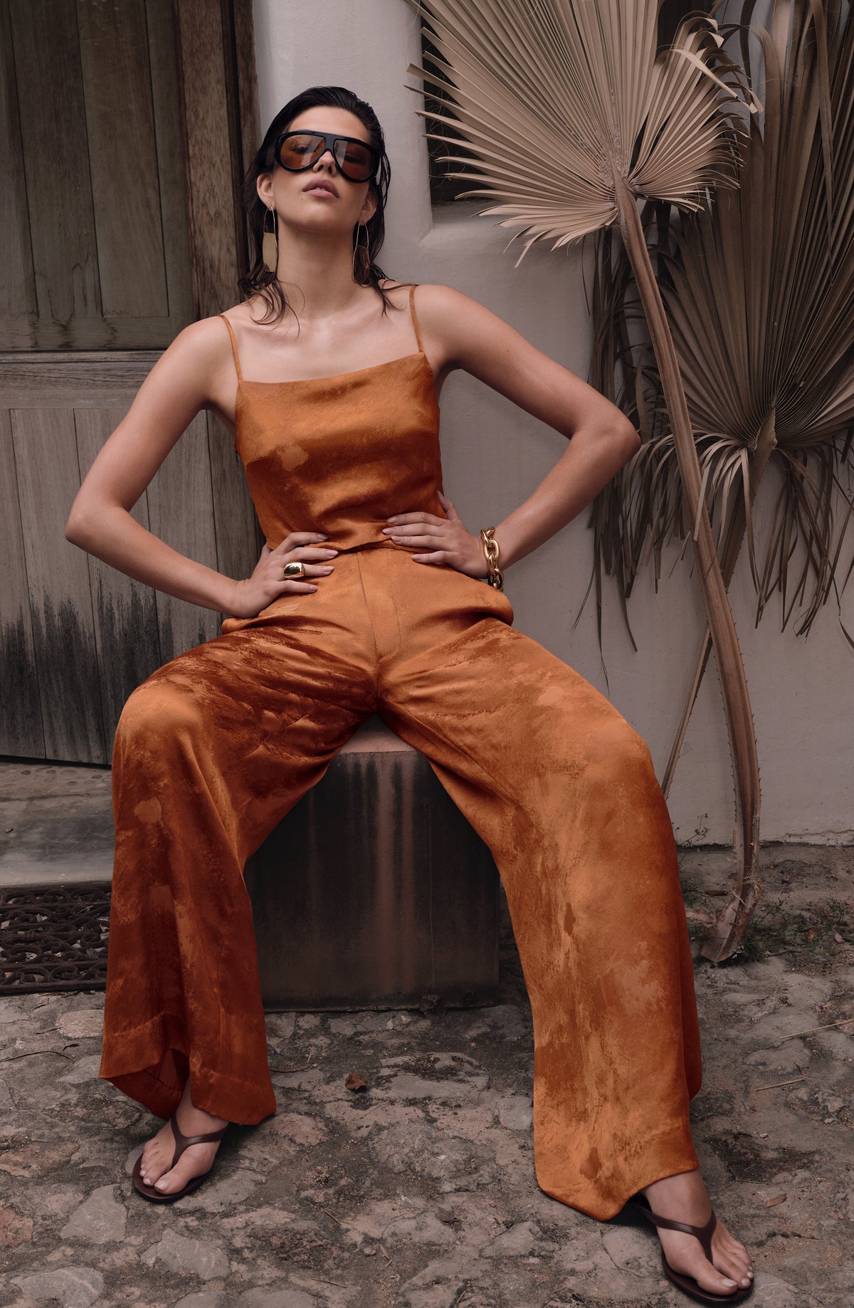 A woman wearing a rust-orange satin jumpsuit and dark sunglasses sits on a metal stool in an outdoor patio setting with a palm leaf in the background.