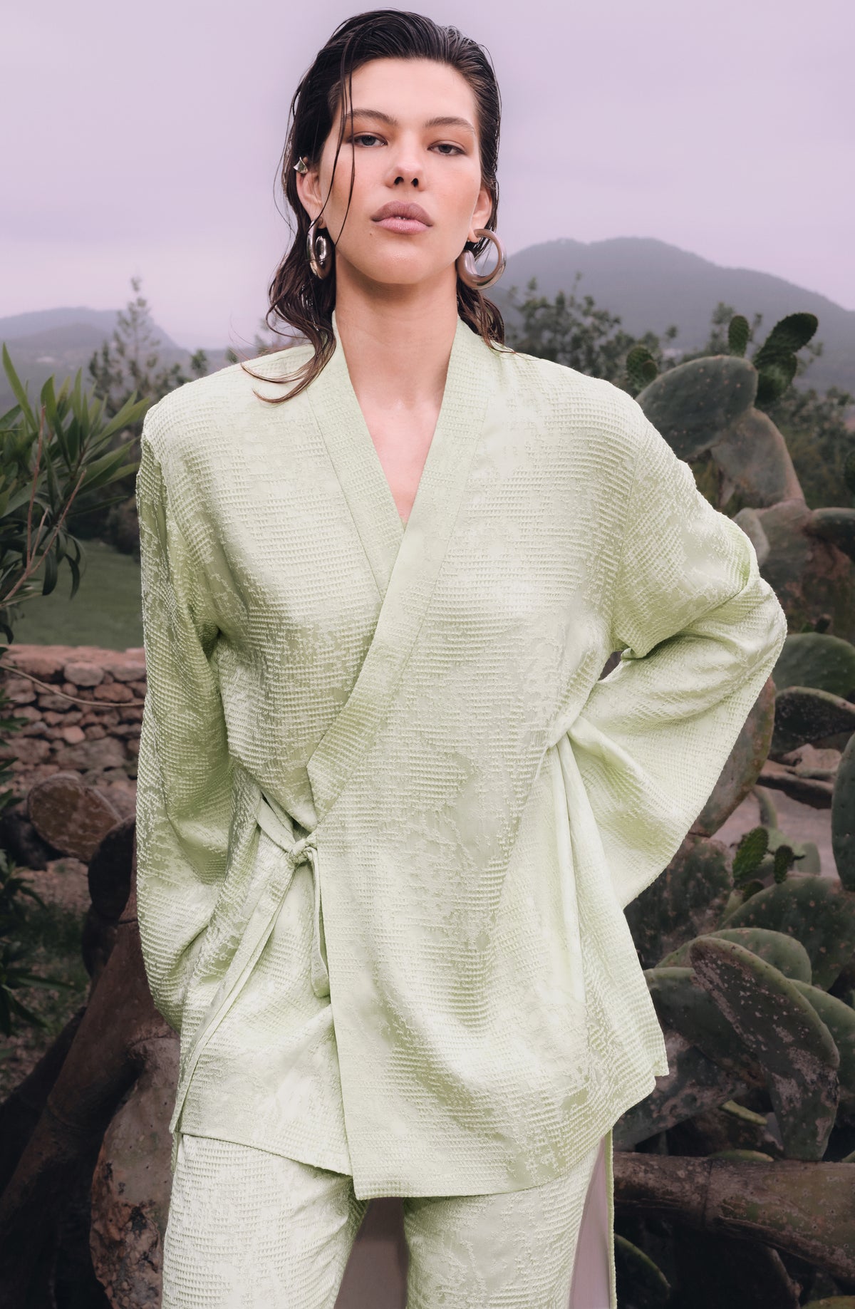 A woman wearing a light sage green textured wrap jacket and matching pants, posed outdoors in front of a scenic background of mountains and cacti.